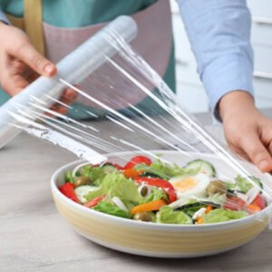 Woman putting plastic food wrap over bowl of fresh salad at wooden table indoors, closeup